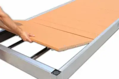 A person places a rectangular, brown decking panel onto a metal frame, assembling an Aluminum Dock from Great Northern Docks.