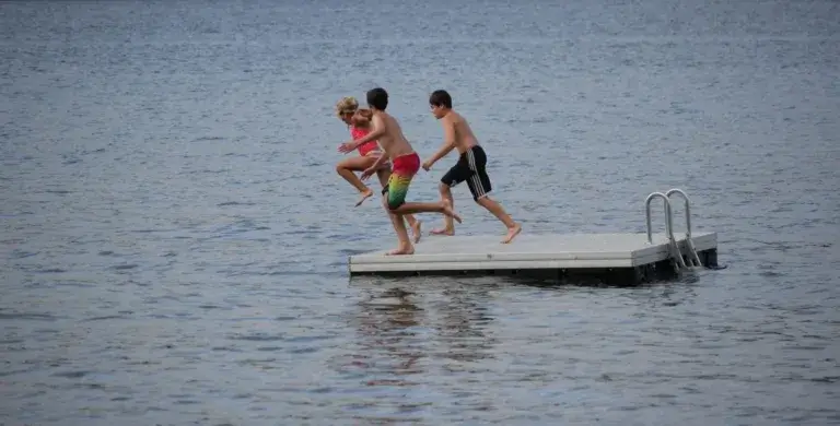 Three children in swimsuits joyfully jump off a floating aluminum dock into a lake, with water rippling around them and a metal ladder on the dock’s edge.