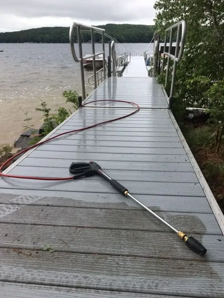 A power washer and hose rest on a wet Maine Docks dock beside a lake, with trees and cloudy skies in the background.
