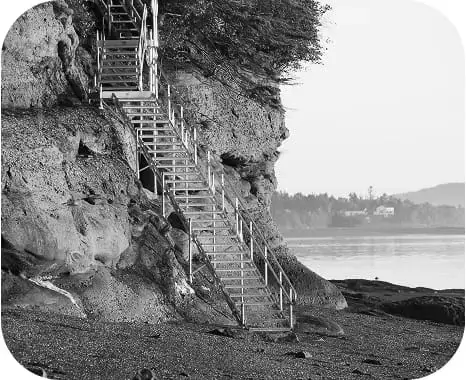 A metal staircase, part of the Great Northern Docks system, leads up a rocky cliff from a pebble beach, with calm water and distant trees in the background.