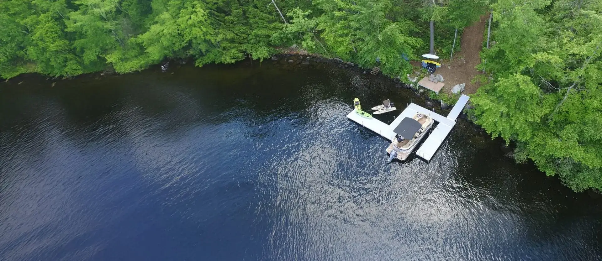 Aerial view of a dock with a boat and kayaks on a calm, dark lake surrounded by dense trees—featuring classic wood docks and a path leading from the shore.