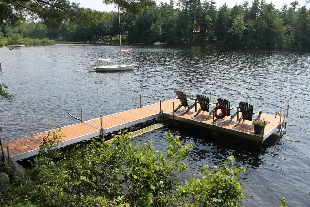 A wooden dock with chairs overlooks a calm lake, surrounded by trees. A sailboat floats nearby, and a lifebuoy hangs from the Maine Docks railing.