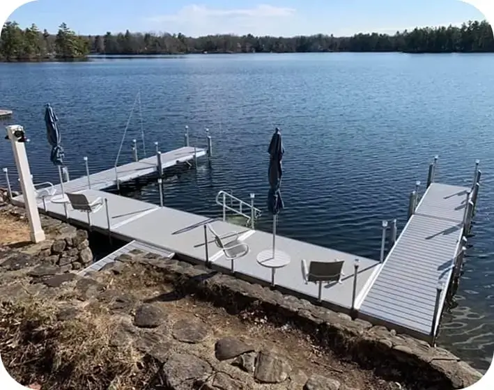 A modern Maine Dock with chairs and closed umbrellas extends over a calm lake, surrounded by trees under a clear sky.