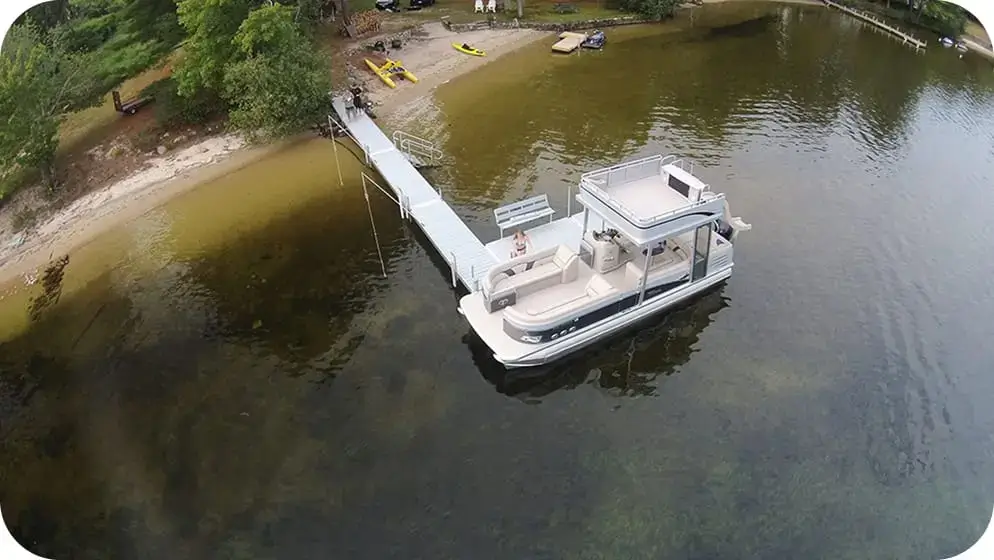 A pontoon boat is docked at a long Great Northern Docks pier extending from a sandy lakeshore with trees, kayaks, and people visible on land.