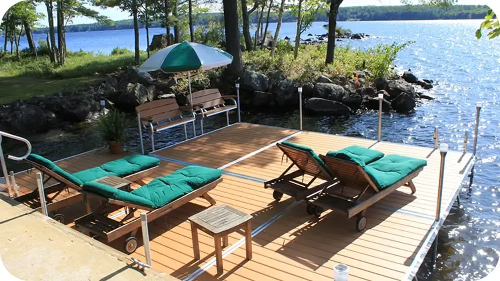 A Great Northern Docks wooden dock extends over the lake, featuring two green-cushioned loungers, a bench with umbrella, small table, and surrounded by trees and rocks.