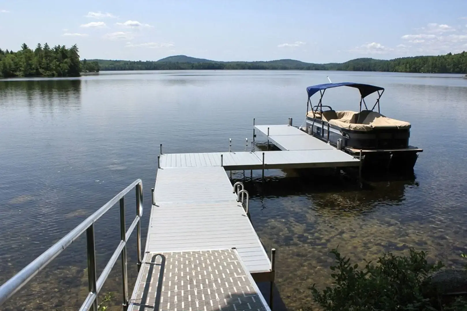 A metal dock by Great Northern Docks extends into a calm lake with a pontoon boat moored at the end; trees and hills surround the water under a blue sky.