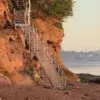 An aluminum dock staircase leads up a rocky cliff beside calm water, with greenery on top and distant hills and buildings across the bay.