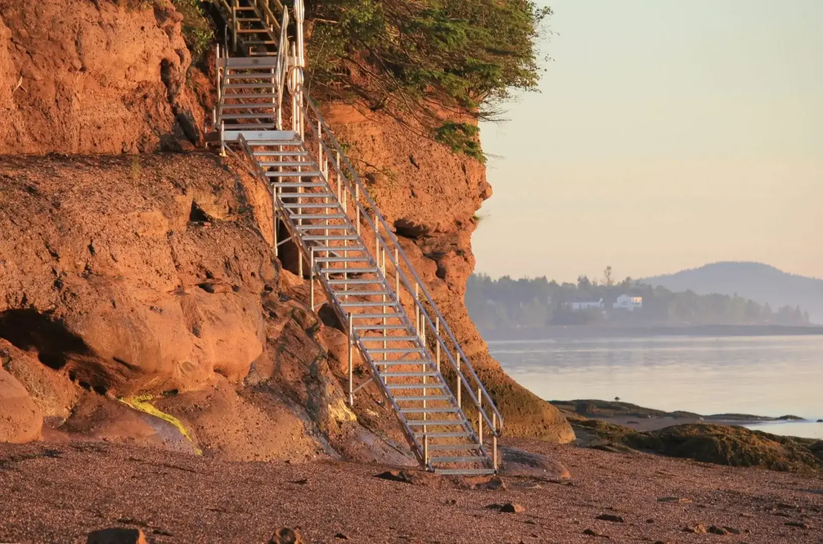 An aluminum dock staircase leads up a rocky cliff beside calm water, with greenery on top and distant hills and buildings across the bay.