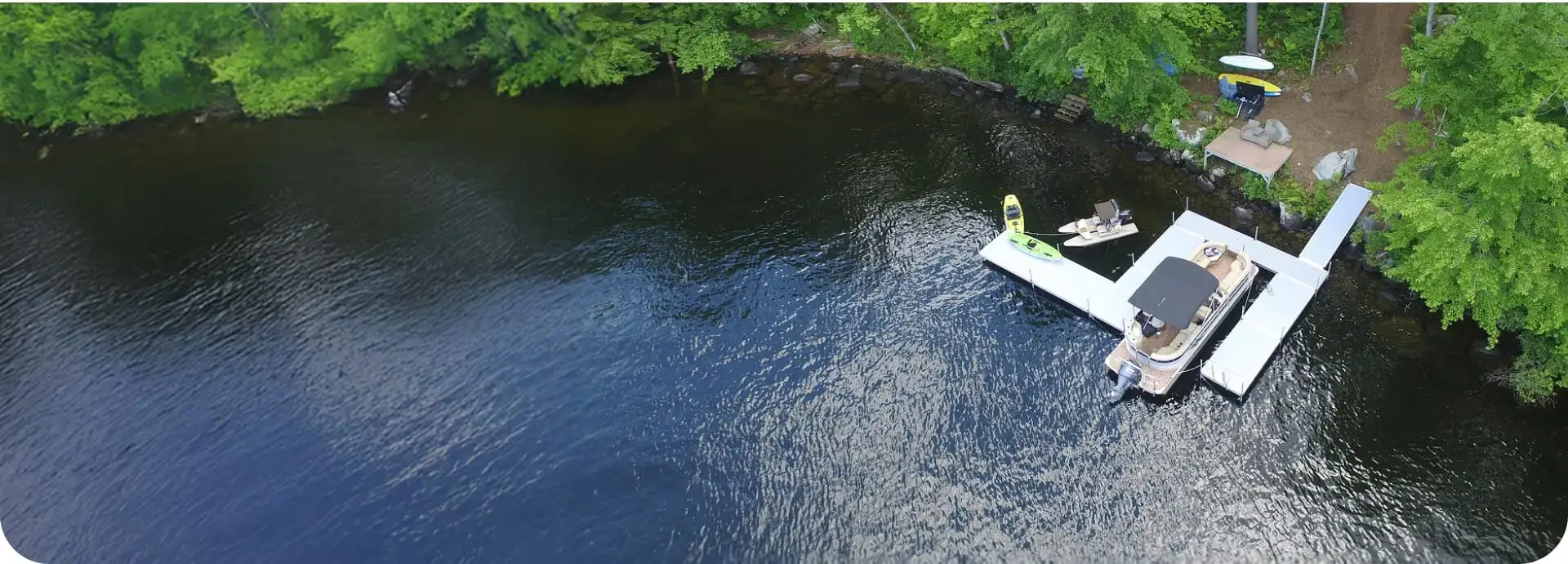 Aerial view of a Wood Dock with a boat and kayaks on a calm lake, surrounded by lush green trees and a small clearing on the shore.