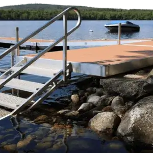 A metal dock by Great Northern Docks with stairs extends into a calm lake surrounded by rocks, boats, and forested hills in the background.