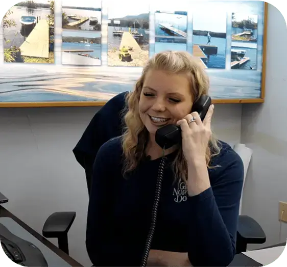 A smiling woman with blonde hair talks on a corded phone at her desk, with Great Northern Docks and lake photos displayed on the wall behind her.