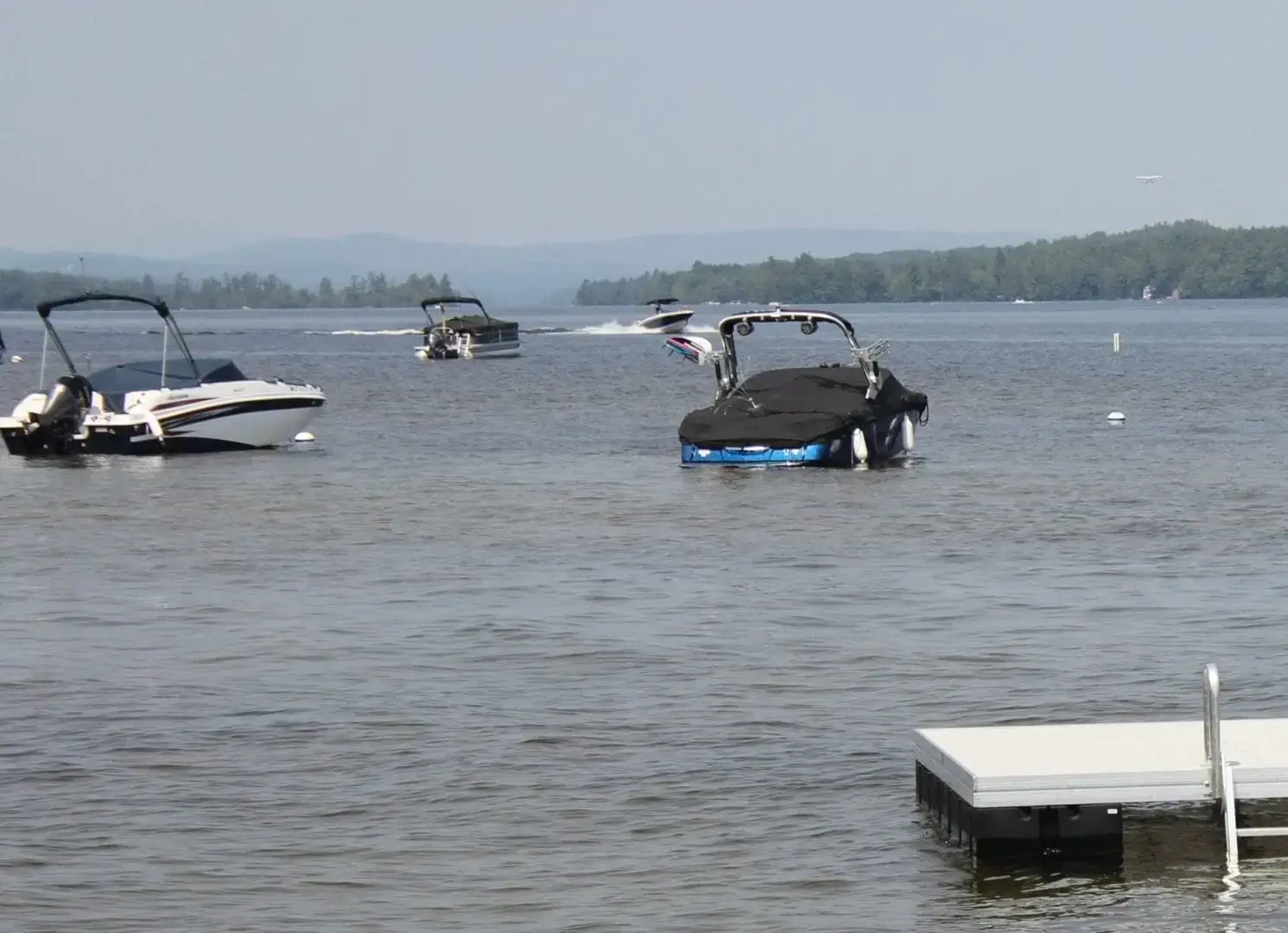 Several boats float on a lake near Maine Docks under a clear sky, secured by Mooring Ball, with wooded hills in the background.
