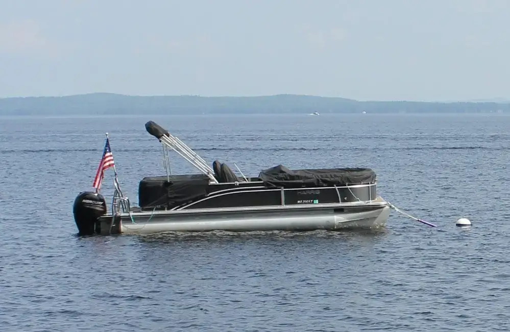 A pontoon boat with a U.S. flag is anchored to a Mooring Ball near Great Northern Docks, with calm water and distant land under a clear sky.