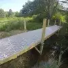 A 4x4 Post Bracket by Great Northern Docks supports a wood and plastic footbridge over a stream, surrounded by grass, trees, and shrubs.
