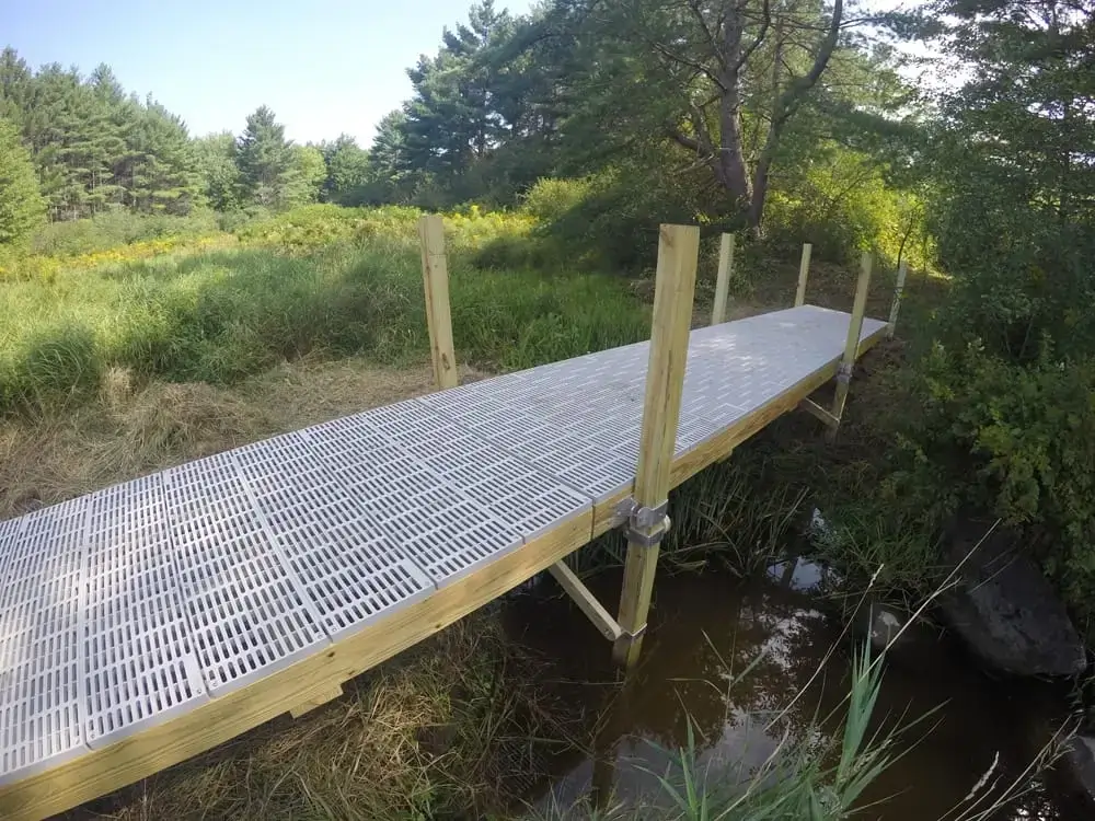 A 4x4 Post Bracket by Great Northern Docks supports a wood and plastic footbridge over a stream, surrounded by grass, trees, and shrubs.