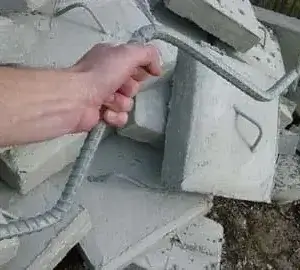 A hand holds a DIY Anchor Rod for Concrete Block in front of broken slabs with metal handles, commonly used near Maine Docks or beside wood docks.