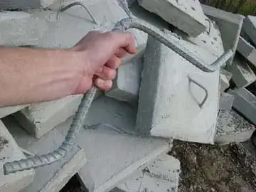 A hand holds a DIY Anchor Rod for Concrete Block in front of broken slabs with metal handles, commonly used near Maine Docks or beside wood docks.
