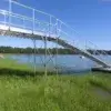 A walkway with railings made from 1.5" Sch. 40 Galvanized Pipe leads to a floating dock on a calm lake, surrounded by grass and trees under a blue sky.