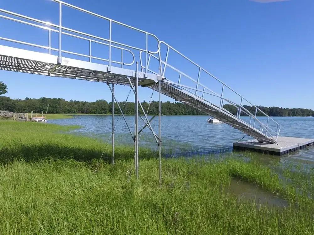 A walkway with railings made from 1.5" Sch. 40 Galvanized Pipe leads to a floating dock on a calm lake, surrounded by grass and trees under a blue sky.