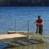 A person uses a Leveling Winch to assemble or repair a wood dock in shallow lake water on a sunny day, surrounded by trees and scenic Maine views.