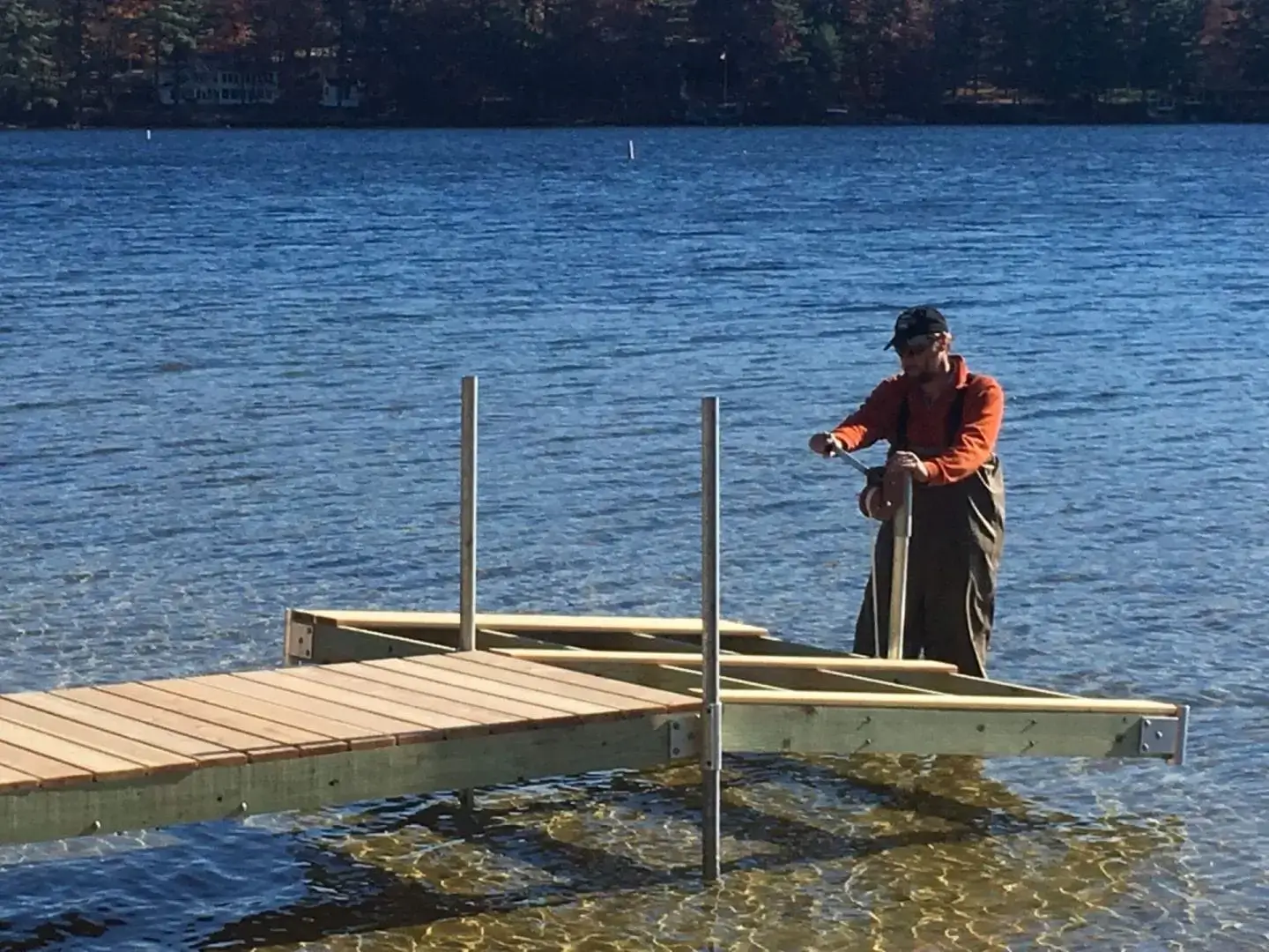 A person uses a Leveling Winch to assemble or repair a wood dock in shallow lake water on a sunny day, surrounded by trees and scenic Maine views.