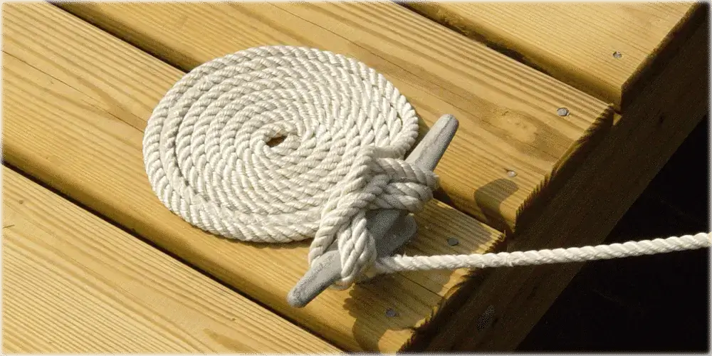 A coiled white rope is neatly placed on a wood dock, with one end tied to a Dock Cleats Galvanized (2 Pack) gray cleat.