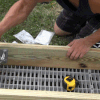Two people are assembling a wooden frame for Maine docks using the Corner Iron 90º bracket, with tools and screws on top and grass underneath; one hammers while the other holds the wood.