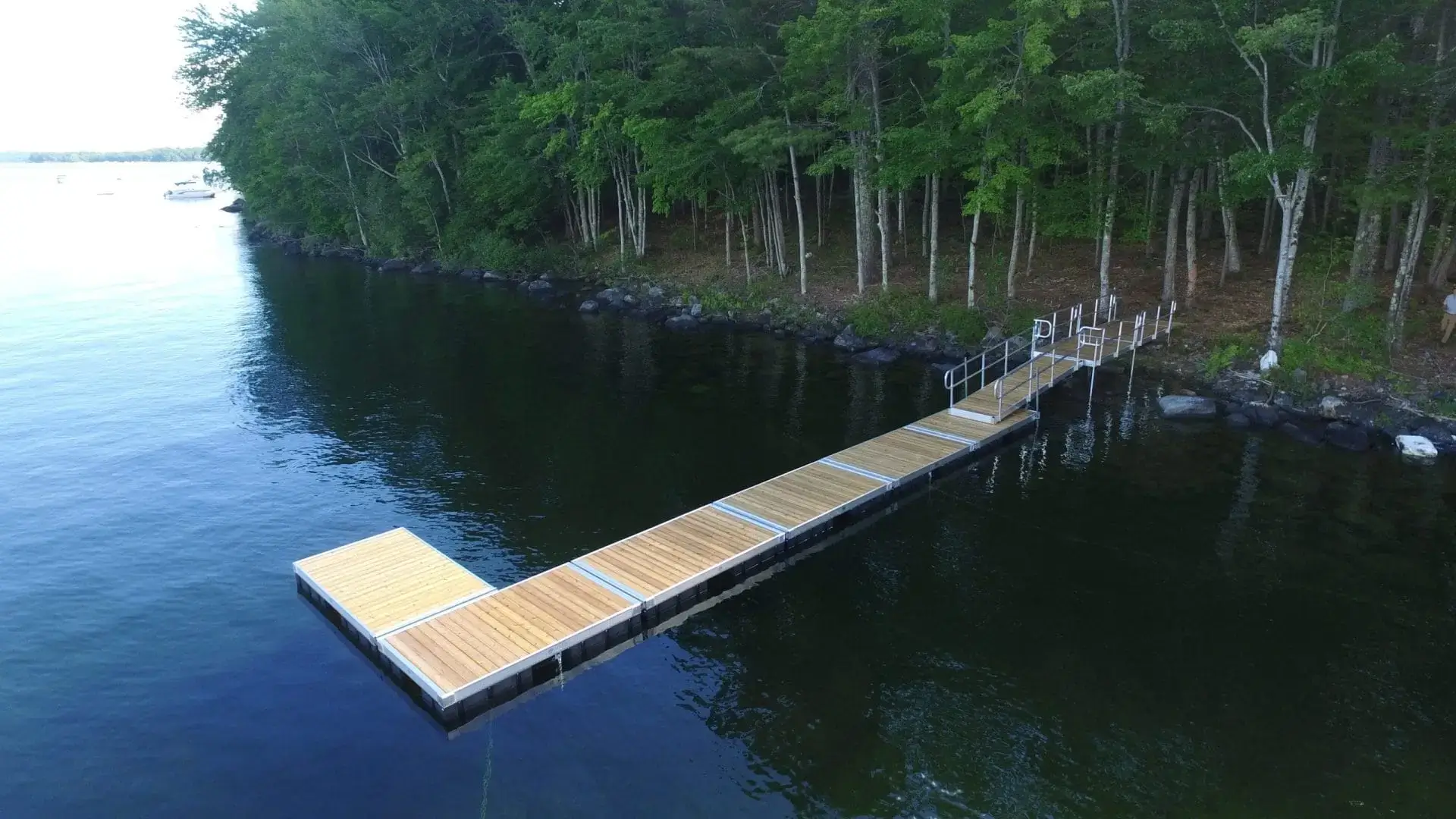 A Floating Dock from Great Northern Docks extends over calm, dark water near a forested shoreline, with boats visible in the distance.