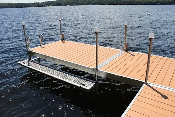 A dock with 1.5" Sch. 40 Galvanized Pipe railings and lights extends over a lake, with grated platform sides and Maine Docks along the tree-lined shore.