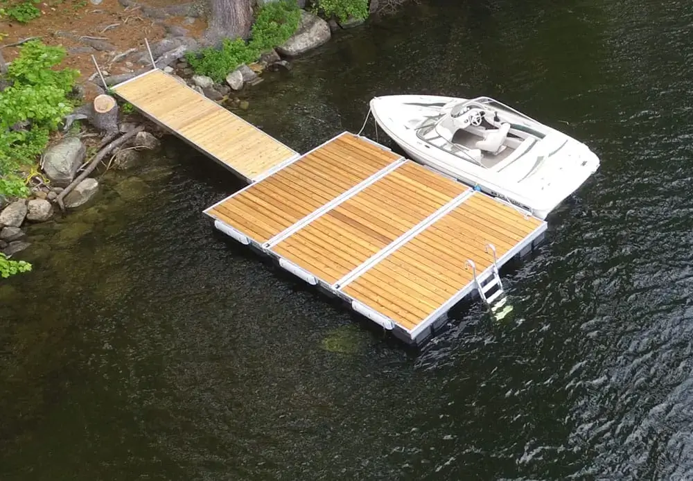An Aluminum Ladder for Dura-LITE Docks is attached to a floating dock on a lake, with green trees and rocks along the shore at Great Northern Docks.