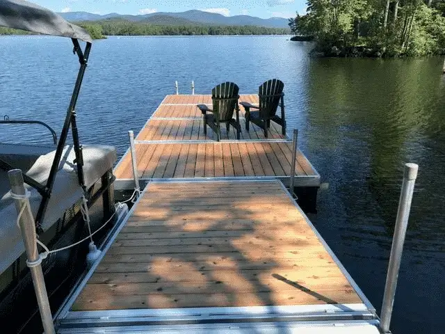 A wooden dock by Great Northern Docks on a lake with two Adirondack chairs, overlooking trees and mountains in the background.