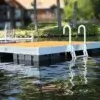 An Aluminum Ladder for Dura-LITE Docks is installed on a floating wood dock with metal railings on a calm lake, trees and a house blurred in the background.