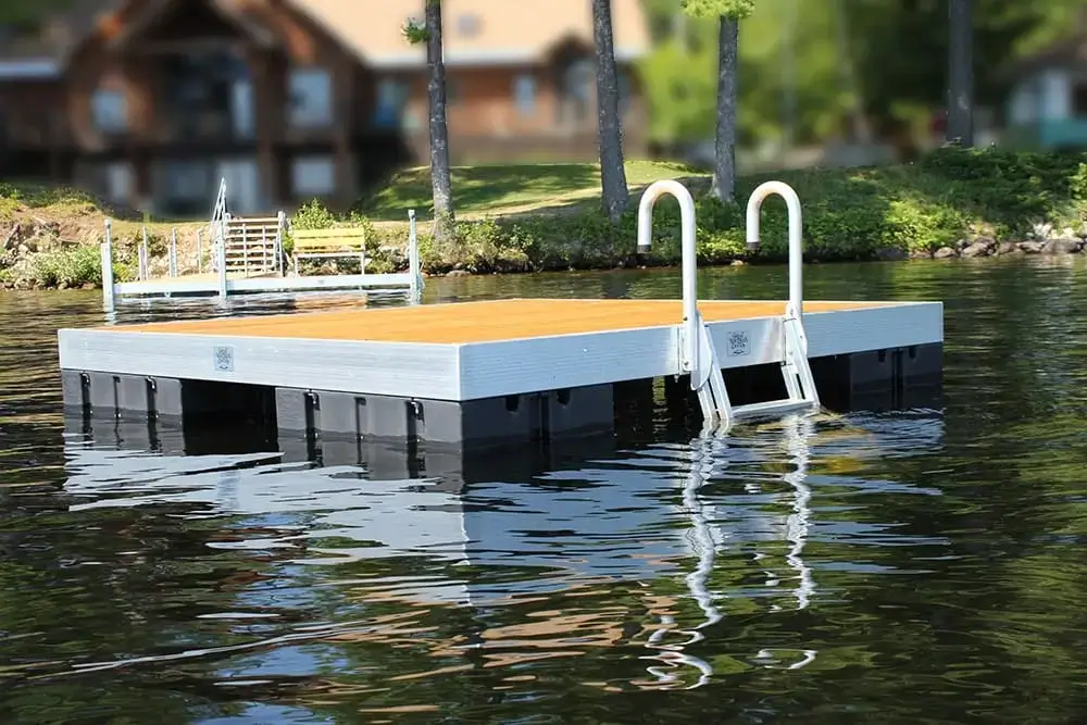 An Aluminum Ladder for Dura-LITE Docks is installed on a floating wood dock with metal railings on a calm lake, trees and a house blurred in the background.