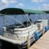 A pontoon boat with a green canopy is secured to a wooden pier using a Mooring Whip (2 Pack) on a lake under a partly cloudy sky.