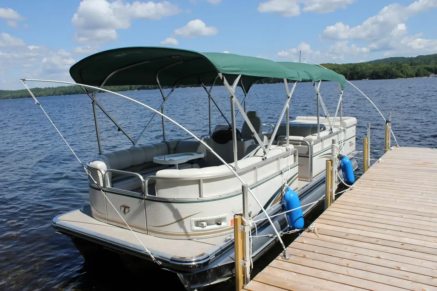 A pontoon boat with a green canopy is secured to a wooden pier using a Mooring Whip (2 Pack) on a lake under a partly cloudy sky.