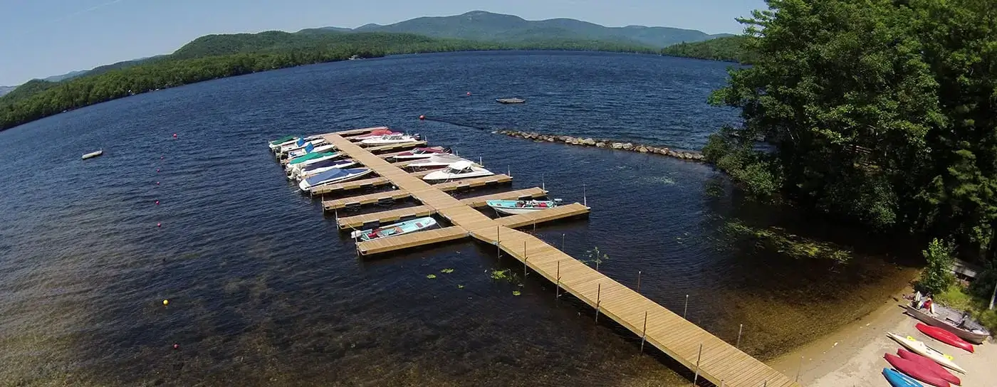 A wooden dock from Great Northern Docks anchors boats on a serene lake, surrounded by green trees, distant mountains, and kayaks resting on a sandy Maine beach.