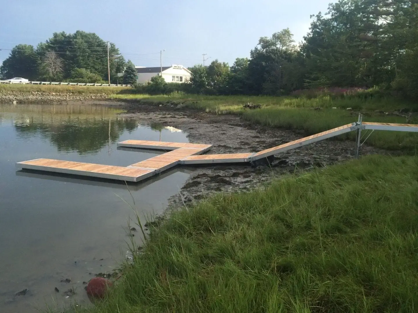 A reinforced hinge plate connects a floating wood dock to grassy land, creating the classic Maine dock setup with trees and a house in the background.