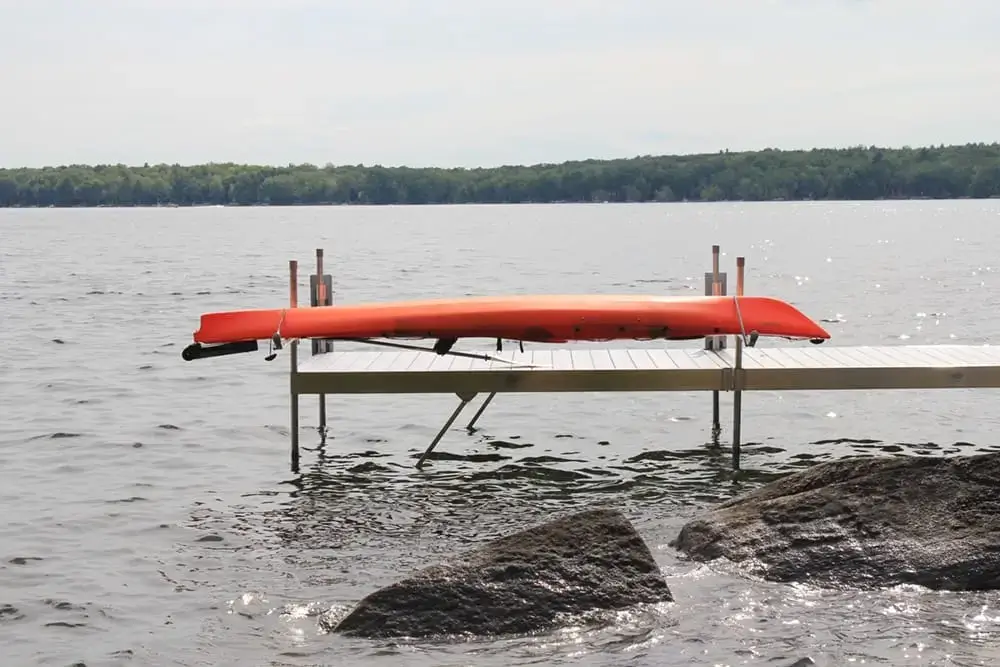 A Kayak & Canoe Arm (Single Arm) secures a red kayak upside down on a wooden dock over a calm lake, framed by rocks and distant tree-lined shores—classic Great Northern Docks.