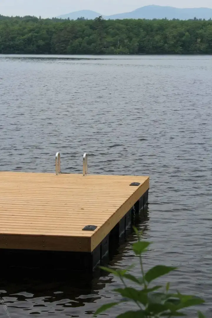 A PermaFloat foam-filled dock with metal ladder extends over a calm lake, featuring classic Maine style with trees and mountains in the background.