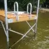 An Aluminum Dock Ladder from Great Northern Docks stands in shallow, clear water by a shoreline with trees in the background.