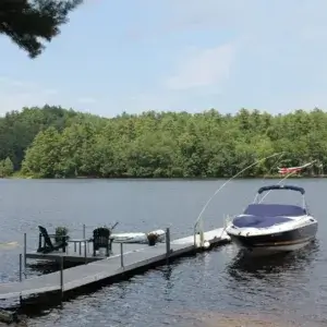 A Fixed Dock on Legs supports two chairs and a potted plant over a calm lake with a moored boat, dense trees across the water, and a partly cloudy sky overhead.