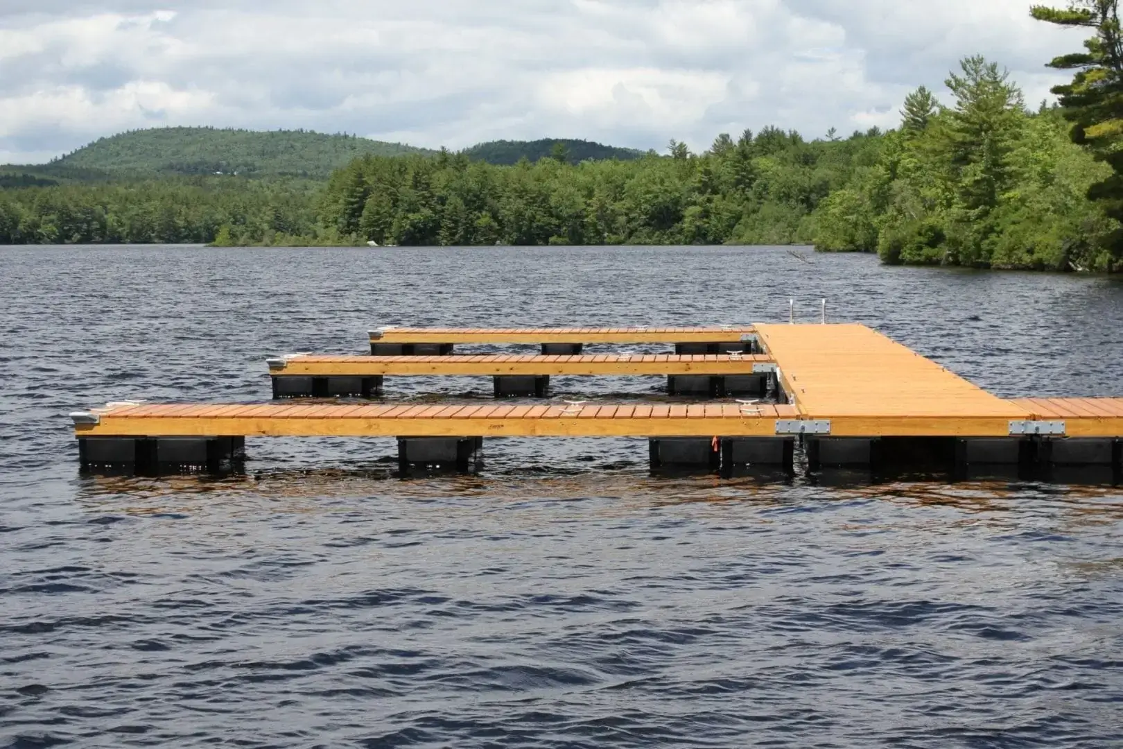 An Anchor Plate Keyhole by Great Northern Docks is installed on a wooden dock extending into a lake, surrounded by trees and hills under a partly cloudy sky.