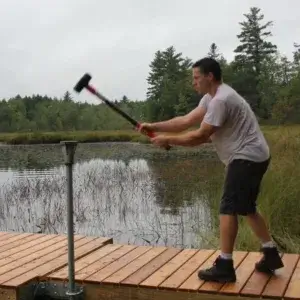 A man in a t-shirt and shorts uses Pipe Driving Caps on a Great Northern Docks wooden dock by a lake, surrounded by trees and tall grass on a cloudy day.