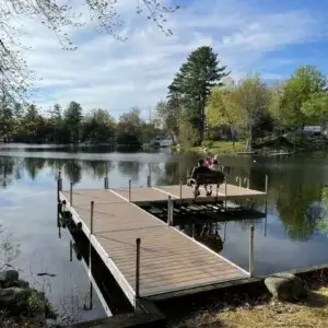Two people sit on a Fixed Dock on Legs, overlooking a calm lake surrounded by trees and houses on a sunny day.