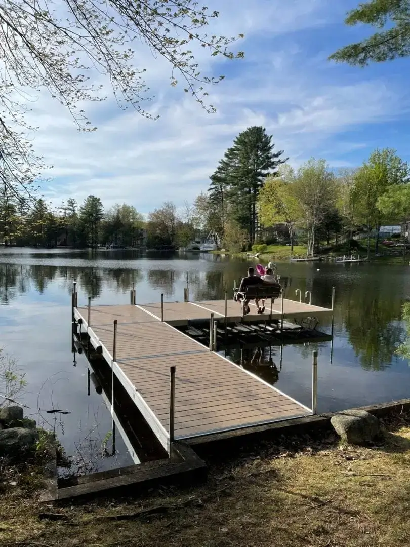Two people sit on a Fixed Dock on Legs, overlooking a calm lake surrounded by trees and houses on a sunny day.