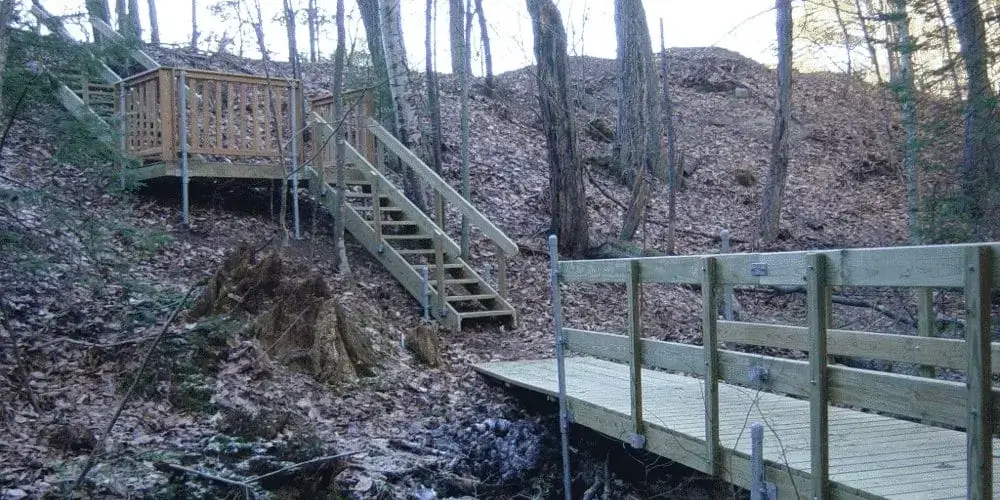 A rail bracket supports a staircase and bridge ascending a leaf-strewn forested hill, surrounded by tall trees, reminiscent of classic Wood Docks.