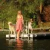 A woman, a young girl, and a golden retriever stand on a Floating Dock over a calm lake with green trees and warm sunlight all around.