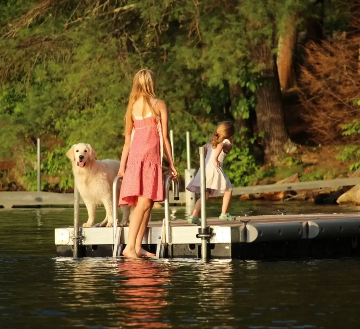 A woman, a young girl, and a golden retriever stand on a Floating Dock over a calm lake with green trees and warm sunlight all around.