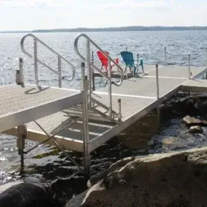 A set of Fully Welded Stairs leads to two colorful chairs on a dock overlooking water, with rocks in front and a distant shoreline.