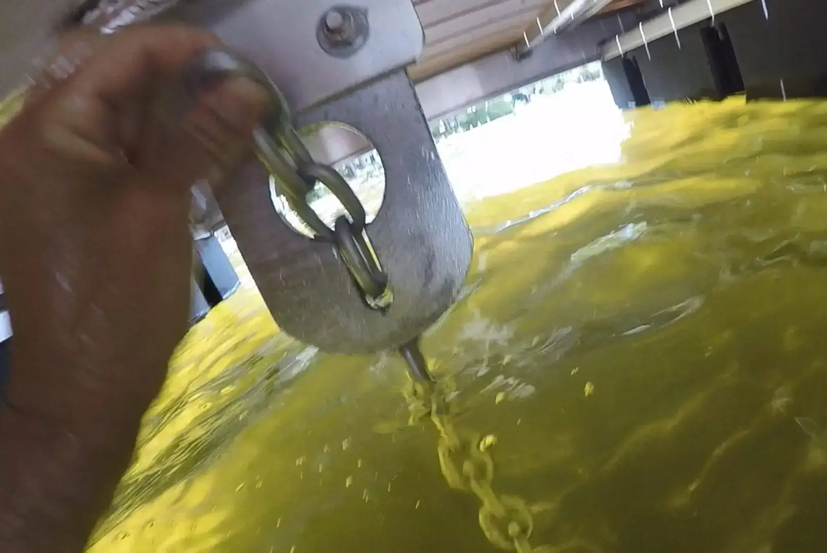 A hand attaches Shackles to a metal chain and large ring above yellow-tinted water, likely beneath sturdy Maine Docks or a classic wood dock platform.